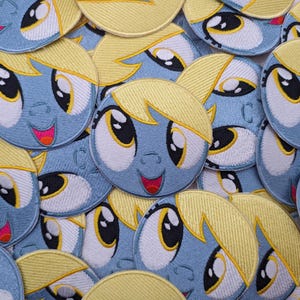 May include: A collection of embroidered patches with a cartoon character's face. The patches are circular, with a light blue face, yellow hair, and large eyes. The character has a smiling mouth with a pink tongue and orange accents.