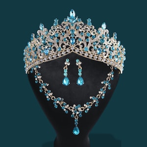May include: A gold-toned tiara, necklace, and earring set with blue and clear crystals. The tiara has a detailed, ornate design. The necklace and earrings feature matching blue teardrop-shaped crystals. The set is displayed on a black mannequin head against a dark teal background.