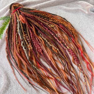 May include: A bundle of colorful dreadlocks in shades of brown, red, and orange. Some dreads are braided, others are adorned with gold accents. The dreadlocks are tied together with a red band.