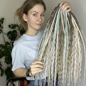 Op de afbeelding: Een bundel lange, golvende synthetische dreadlocks in tinten blond, bruin en lichtblauw. De dreadlocks worden omhoog gehouden, waarbij de textuur en kleurvariaties worden getoond. De stijl lijkt een haarverlenging te zijn.