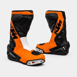 Könnte beinhalten: Paar orangefarbene und schwarze Motorradstiefel. Die Stiefel haben ein hoch geschnittenes Design mit einem weißen, gepolsterten Bereich auf der Vorderseite. Die Stiefel haben die Buchstaben "KM" in Schwarz auf der Seite. Die Stiefel haben eine schwarze Sohle und Zehenbereich.