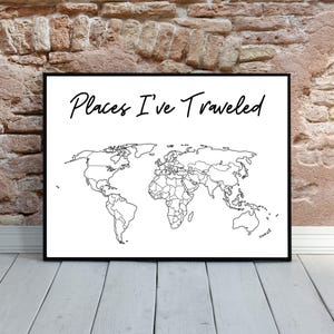 May include: A framed world map print with the text "Places I've Traveled" in a handwritten style. The map is a black and white outline of the continents and countries. The frame is black, and the background is a brick wall and wooden floor.