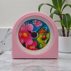 May include: A pink, arched-shaped analog clock with a floral design on the face. The clock face features a colorful illustration of flowers, a house, and a ladybug. The clock has black hands and white numbers.