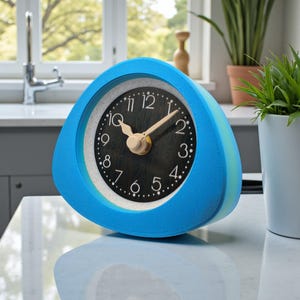 May include: A blue triangular alarm clock with a black clock face and white numbers. The clock hands are light brown. The clock is sitting on a white surface. The clock is a modern design.