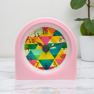 May include: A pink, arched-top alarm clock with a colorful geometric face. The clock face features a pattern of triangles in various colors, including yellow, pink, green, and blue. The clock hands are black, and the numbers are in black. The clock is approximately 4 inches tall.