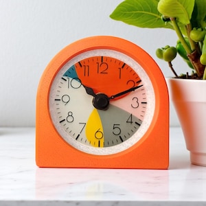 May include: An orange, semi-circular alarm clock with a colorful face. The clock face features a segmented design with orange, white, yellow, gray, and blue sections. Black hands point to the time. A small plant in a white pot is in the background.