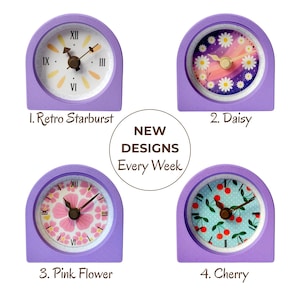 May include: Four small, lavender-colored clocks with different face designs. The clocks include a retro starburst, daisy, pink flower, and cherry design. The text "NEW DESIGNS Every Week" is also present.