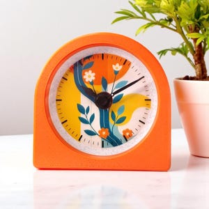 May include: An orange, arched-shaped alarm clock with a floral design on the clock face. The clock face features a tree with flowers and leaves in shades of yellow, orange, and blue. The clock hands are black.