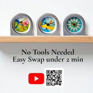 Op de afbeelding: Drie kleine, zilverkleurige klokken met kleurrijke bloemen- en vlinderontwerpen op een houten plank. De wijzerplaten tonen een kolibrie, een vlinder en een madeliefje. De tekst "No Tools Needed Easy Swap under 2 min" staat eronder.