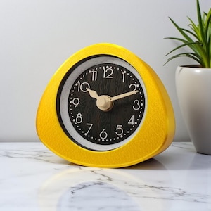 May include: A yellow triangular alarm clock with a black face and white numbers. The clock has a gold-colored frame and hands. The clock is sitting on a marble surface.