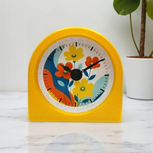 May include: A yellow, retro-style clock with a floral design on the face. The clock face features yellow and orange flowers against a blue and white background. The clock has black hands and a textured yellow frame.