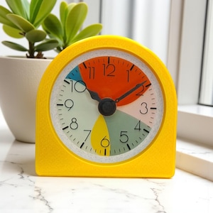 May include: A yellow retro-style alarm clock with a colorful face. The clock face features orange, blue, green, and yellow sections. The clock has black hands and a black knob. A small potted plant is in the background.