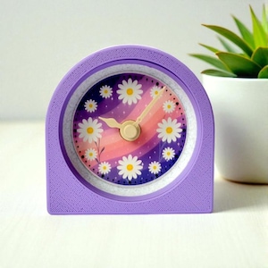 May include: A lavender-colored, arched desk clock with a daisy-themed face. The clock face features a gradient of pink, purple, and white, with white daisies. The clock hands and center are a light beige color. A small succulent plant in a white pot is in the background.
