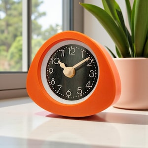 May include: An orange triangular alarm clock with a black face and white dotted hour markers. The clock has beige hands and a beige knob. The clock is sitting on a white surface, with a window and a potted plant in the background.