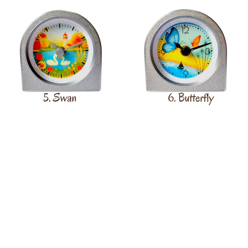 Op de afbeelding: Twee kleine, zilverkleurige klokken met gewelfde bovenkanten. E&eacute;n wijzerplaat toont zwanen op een meer, de andere een vlinder. De tekst "5. Swan" en "6. Butterfly" staat eronder. De wijzerplaten hebben een kleurrijk, geschilderd ontwerp.