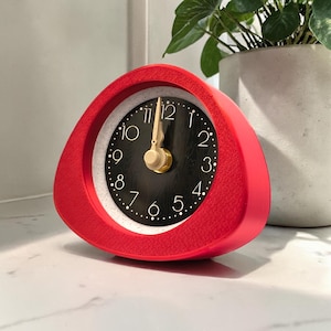 May include: A red, triangular-shaped alarm clock with a black clock face and gold-colored hands. The clock has white dots marking the minutes and gold numerals. The clock is on a white surface.