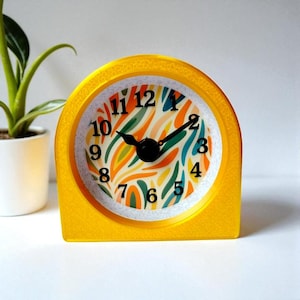 May include: A bright yellow retro-style alarm clock with a colorful face featuring orange, green, and blue abstract designs. The clock has black hands and numbers, and a white background. A small plant in a white pot is in the background.