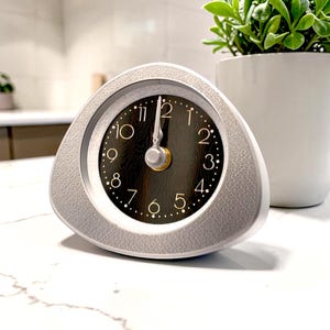 May include: A triangular-shaped analog clock with a textured light gray frame and a dark brown wood-grain patterned face. The clock features gold-colored numerals from one to twelve, a circular minute track, and silver-colored hour and minute hands.