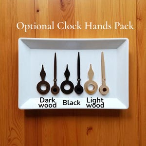 Op de afbeelding: Een witte rechthoekige lade toont vijf wijzers in donker hout, bruin, zwart en licht hout. De tekst "Optional Clock Hands Pack" staat boven de wijzers en de woorden "Dark wood", "Black" en "Light wood" staan eronder.