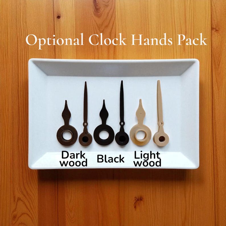 Op de afbeelding: Een witte rechthoekige lade toont vijf wijzers in donker hout, bruin, zwart en licht hout. De tekst "Optional Clock Hands Pack" staat boven de wijzers en de woorden "Dark wood", "Black" en "Light wood" staan eronder.