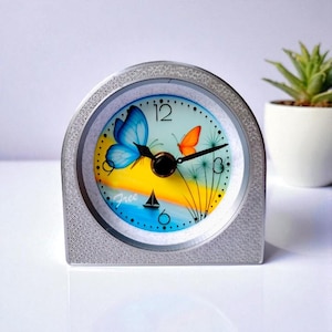 May include: Silver-toned, arched desk clock with a colorful face featuring a blue butterfly, an orange butterfly, and the word "Free" with a sailboat. The clock has black hands and a black clock face.
