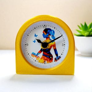May include: A yellow, arched desk clock with a white face featuring a silhouette of a person lifting weights. The person is depicted with blue and orange hues. Two blue butterflies are on the clock face. The clock hands are black.