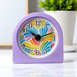 May include: A lavender-colored, arched desk clock with a colorful, abstract face. The clock face features a rainbow of colors and black hands. The clock is set against a blurred background with a plant.
