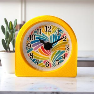 May include: A bright yellow, arched desk clock with a colorful, abstract floral design on the clock face. The clock hands are black. A small cactus in a white pot is to the left of the clock.
