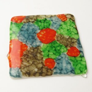 May include: A colorful abstract design on a square coaster with a drip of glaze on the bottom right corner. The design features shades of green, blue, orange, and brown.