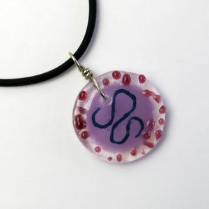 May include: A handmade pendant necklace featuring a round, translucent purple charm with a dark blue swirl design. The charm is accented with red dots around the edge and hangs from a black cord.