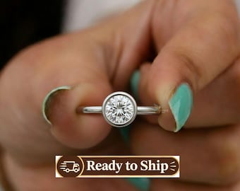 May include: A silver-toned ring featuring a single round, brilliant-cut clear stone set in a smooth bezel setting. The band is thin and polished, showcasing a minimalist design that highlights the central gemstone's facets and clarity.