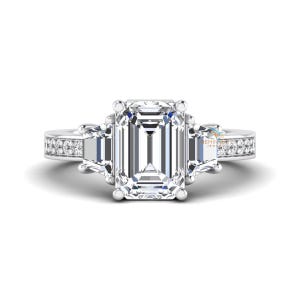 May include: A silver engagement ring with a large emerald-cut center stone flanked by two smaller trapezoid-cut stones. The band is adorned with small, round accent stones. The ring is set against a white background.