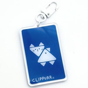 May include: A rectangular keychain with a blue background featuring two white cat-shaped designs. The keychain has a white metal clip and the word "CLIPPURR®" is printed at the bottom.