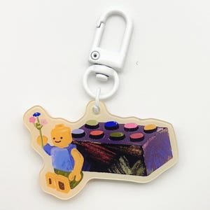 May include: A clear acrylic keychain featuring a colorful illustration of a Lego-style figure holding a flower next to a purple block with colorful studs. The keychain has a white metal clasp.
