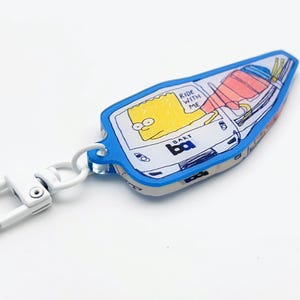 May include: A blue keychain featuring a cartoon image of Bart Simpson riding a vehicle. The keychain has a white clasp and the text "Ride with me" and "Bart" are visible. The design is outlined in black.