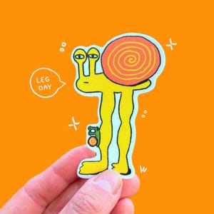 May include: A whimsical sticker featuring a yellow snail with long legs, a spiral shell, and the text "LEG DAY" in a speech bubble. The snail is set against an orange background with white accents.