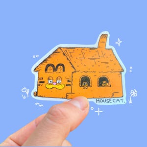 May include: Orange sticker depicting a cartoon house with a cat face and the text "HOUSE CAT." The house has two arched windows and two smaller windows with cat faces. The sticker is held by a hand against a blue background.