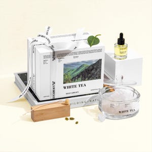 May include: A white gift box set with the words "WHITE TEA" and "ELLE CADEAUX" on the front. The set includes a small bottle of oil, a glass jar with crystals, and a wooden block with a small wooden spoon. A white ribbon is tied around the box.