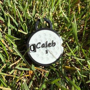 May include: A white and black golf ball-shaped tag with the name "Caleb" and the number "3" printed on it. The tag has a black loop at the top and is set against a background of green grass.
