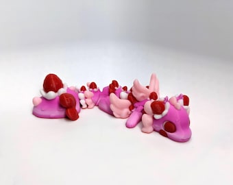 May include: A whimsical, handmade sculpture of a pink creature with red and white accents. The creature has a long body, wings, and strawberry-like details. The sculpture is set against a white background.