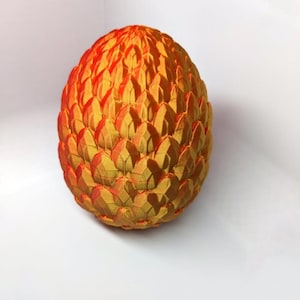 May include: A decorative, egg-shaped object with a textured surface. The egg is primarily yellow with orange highlights, creating a gradient effect. The surface is composed of overlapping, scale-like segments.