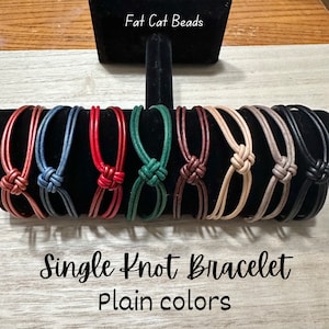 May include: Eight leather bracelets with a single knot in various colors, including red, green, brown, beige, black, and blue. The bracelets are displayed on a black stand and the text "Single Knot Bracelet Plain colors" is below the bracelets.