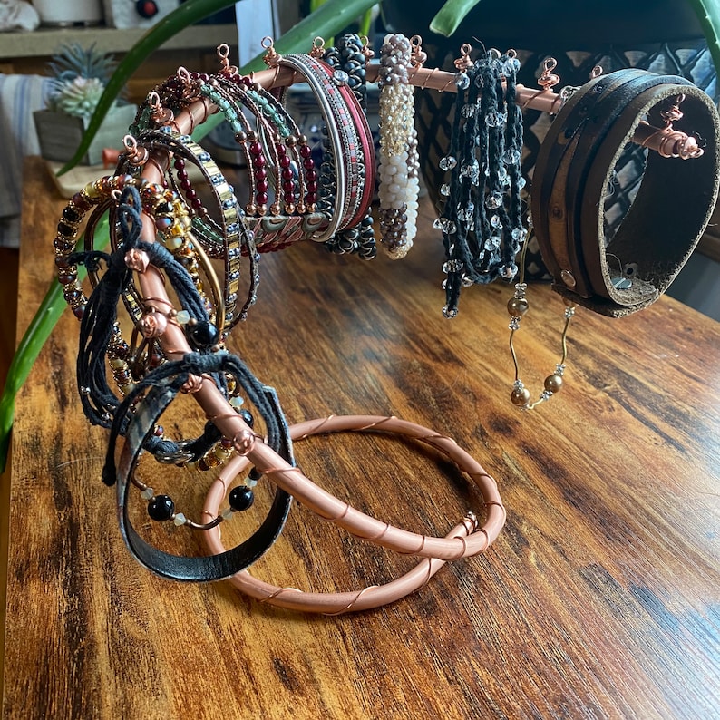 May include: A copper wire jewelry stand with a variety of bracelets and bangles. The stand is shaped like a twisted branch with a circular base. The bracelets are made of leather, beads, and metal.