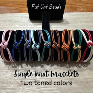 May include: A collection of 10 single knot bracelets with two-toned colors. The bracelets are made of leather and are displayed on a black stand. The text "Fat Cat Beads" and "Single knot bracelets Two toned colors" is visible in the image.