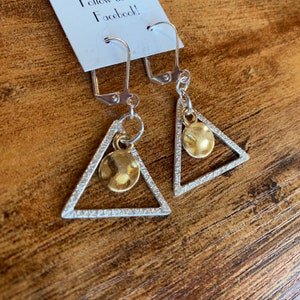 May include: Silver-tone triangle earrings with a hammered gold-tone disc charm. The earrings have lever-back closures and are displayed on a wooden surface. The text "Follow us Facebook!" is visible on a white tag.