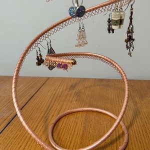 May include: A copper wire earring holder in the shape of a spiral. The holder has multiple loops for hanging earrings. The holder is on a wooden surface and has several pairs of earrings hanging from it.