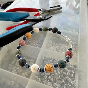 May include: A close-up of a lava rock beaded bracelet with a silver clasp. The bracelet is made with various colors of lava rock beads, including black, white, green, and orange. The bracelet is lying on a white surface with a variety of small silver beads in the background.