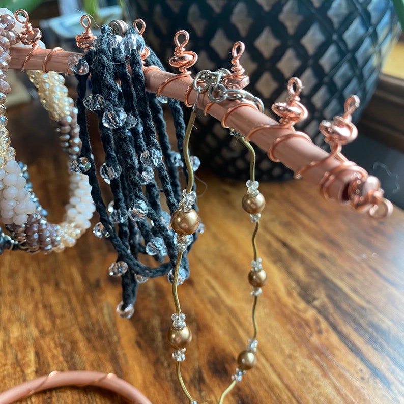 May include: A copper jewelry stand with multiple bracelets and necklaces hanging from it. The stand has a copper wire base and a copper wire top with decorative copper wire swirls. The bracelets and necklaces are made of different materials, including beads, string, and metal.