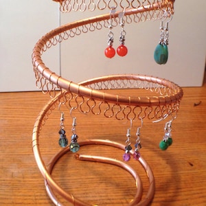 May include: A copper wire earring stand with multiple tiers. The stand is in the shape of a spiral and has several pairs of earrings hanging from it. The earrings are made of various materials, including glass, metal, and beads. The earrings are in a variety of colors, including red, green, blue, and purple.