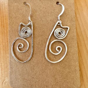 May include: A pair of silver cat-shaped earrings with a spiral design. The earrings are made of wire and have a simple, elegant look.